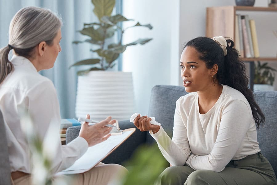 A young woman sits on a couch, holding a tissue and talking to an older woman taking notes, suggesting an anxiety treatment session at Knoll Creek West in IL, in a comfortable, well-lit room with plants.