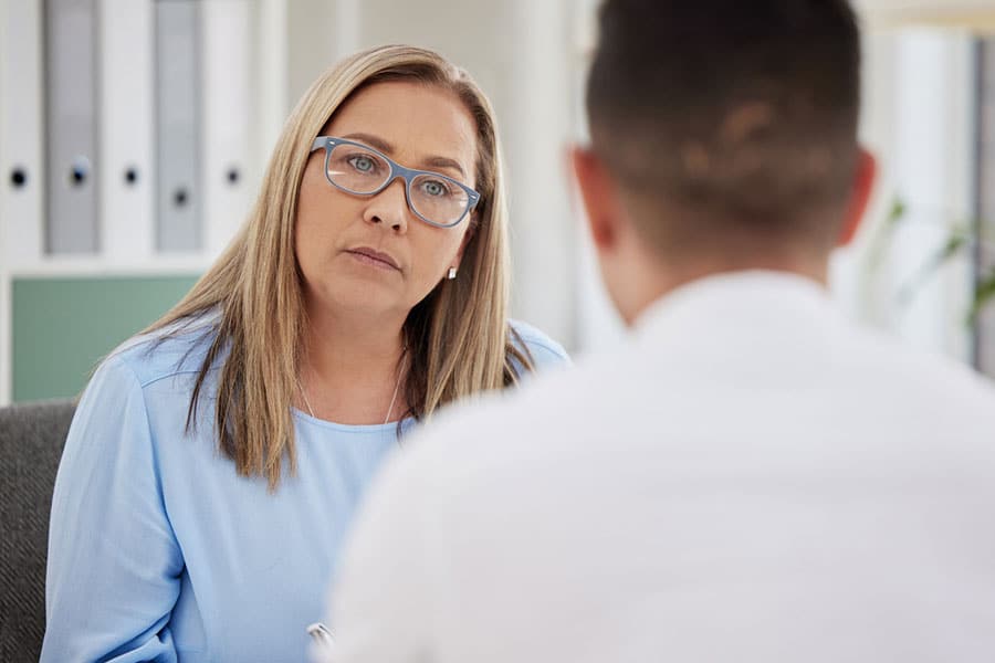 A woman with long blonde hair and glasses listens attentively to a man during a conversation about depression treatment in an office setting. She wears a light blue blouse and appears focused and thoughtful at Knoll Creek West, IL.