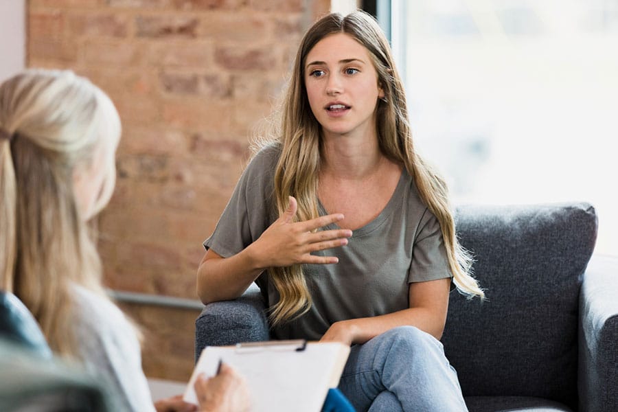 A young woman with long blonde hair sits in a chair, gesturing while talking to a therapist, who is seen from the back holding a notepad. The setting is a room with an exposed brick wall, creating a calm atmosphere ideal for Anxiety Treatment in Wayne IL.