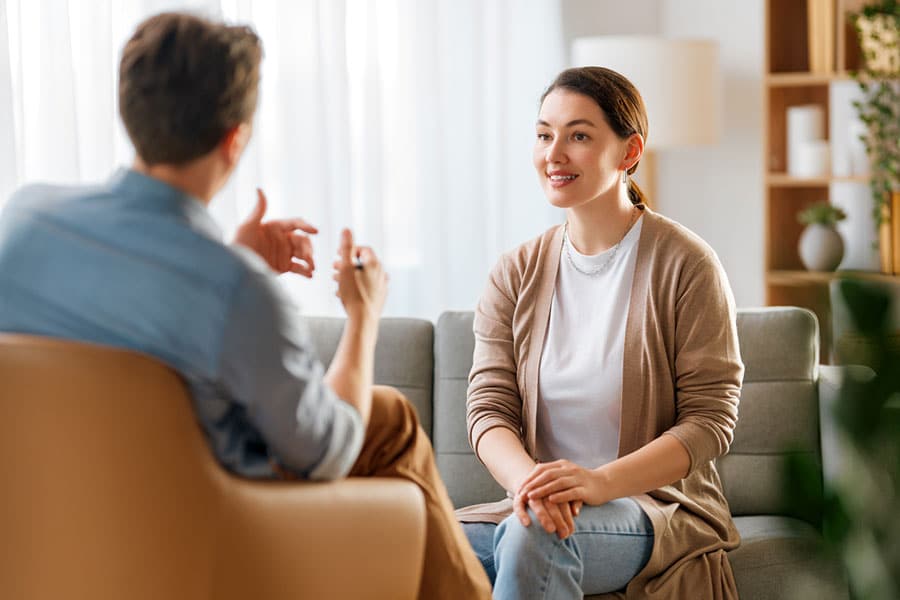 A woman sits on a couch smiling and talking to a man who sits across from her in an armchair. The room, located in West Chicago, IL, is bright with natural light and decorated with plants and shelves—creating a welcoming space for anxiety treatment.