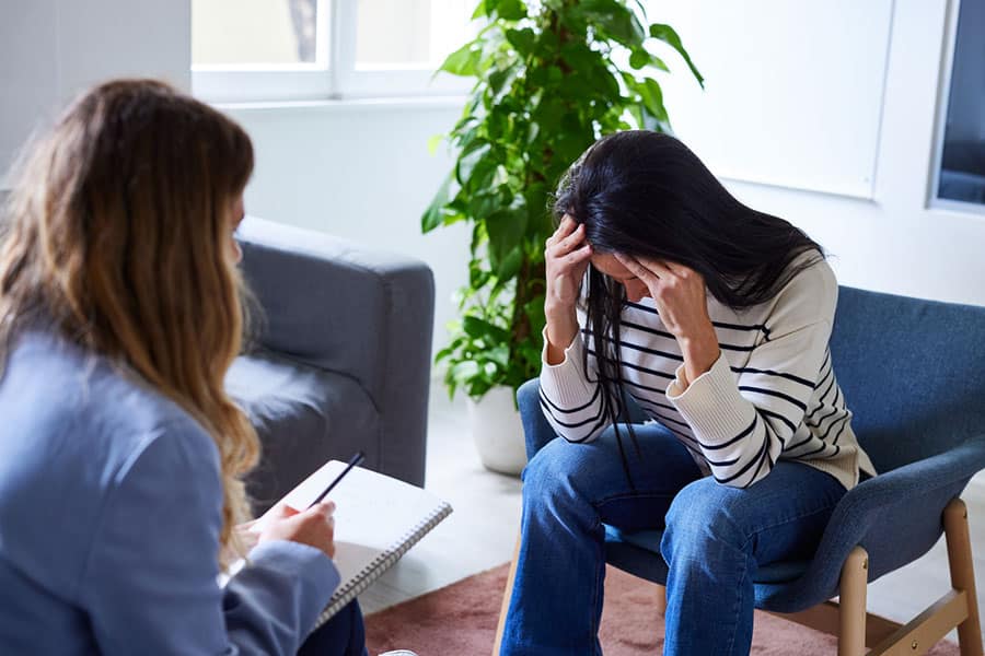 A person sits with their head in their hands, looking distressed, while another person with a notepad listens attentively, suggesting an anxiety treatment session in Wildrose, IL.