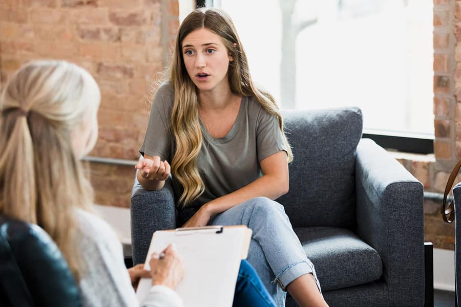 A young woman sits on a gray armchair, discussing depression treatment with a therapist who holds a clipboard. The setting appears to be a cozy office in Wayne, IL, with brick walls and large windows.