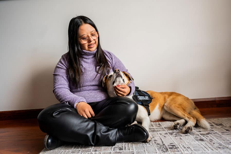A person sitting on a rug and petting a service dog wearing a vest labeled Perro de Servicio during an animal-assisted therapy session.